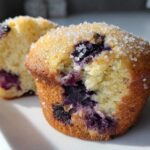 Close-up of two Moist and Fluffy Bakery Style Blueberry Muffins, one cut open to show the crumb and blueberries, topped with coarse sugar.