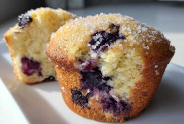 Close-up of two Moist and Fluffy Bakery Style Blueberry Muffins, one cut open to show the crumb and blueberries, topped with coarse sugar.