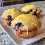 Three moist Lemon Blueberry Muffins topped with a thick lemon glaze, sitting on a white rectangular plate.