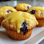Four moist Lemon Blueberry Muffins topped with a thick, bright yellow lemon glaze, sitting on a white plate.
