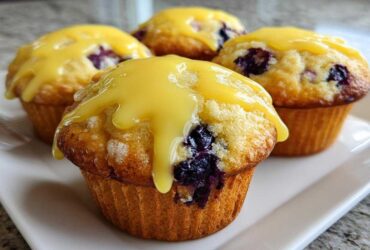 Four moist Lemon Blueberry Muffins topped with a thick, bright yellow lemon glaze, sitting on a white plate.