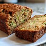 A close-up of a freshly baked, moist Zucchini Bread loaf with one thick slice cut and resting beside it on a white plate.