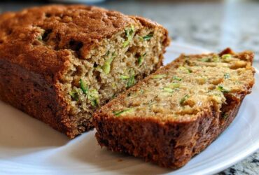 A close-up of a freshly baked, moist Zucchini Bread loaf with one thick slice cut and resting beside it on a white plate.