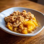 A close-up serving of warm Peach Crumble featuring bright yellow peaches and a golden, crunchy topping.