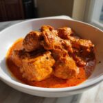 Close-up of tender chicken pieces smothered in vibrant orange, creamy Butter Chicken sauce, served in a white bowl.