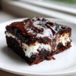 A close-up of a slice of Rich Marshmallow Chocolate Poke Cake showing layers of dark chocolate cake, white marshmallow filling, and glossy chocolate glaze.