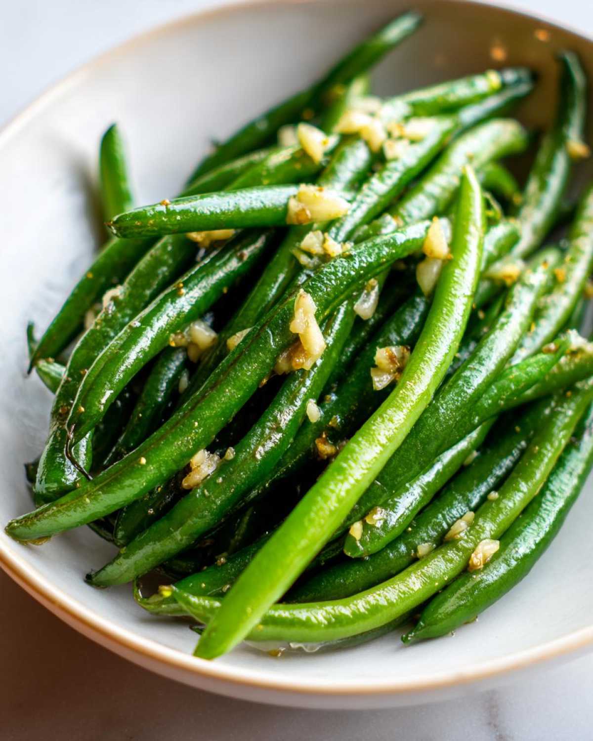 Close-up of vibrant, glossy Roasted Garlic Green Beans topped with minced garlic, served in a light-colored bowl.