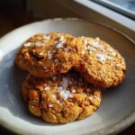 Three golden brown Salted Honey Cookies stacked slightly on a light gray plate, topped with flaky sea salt.