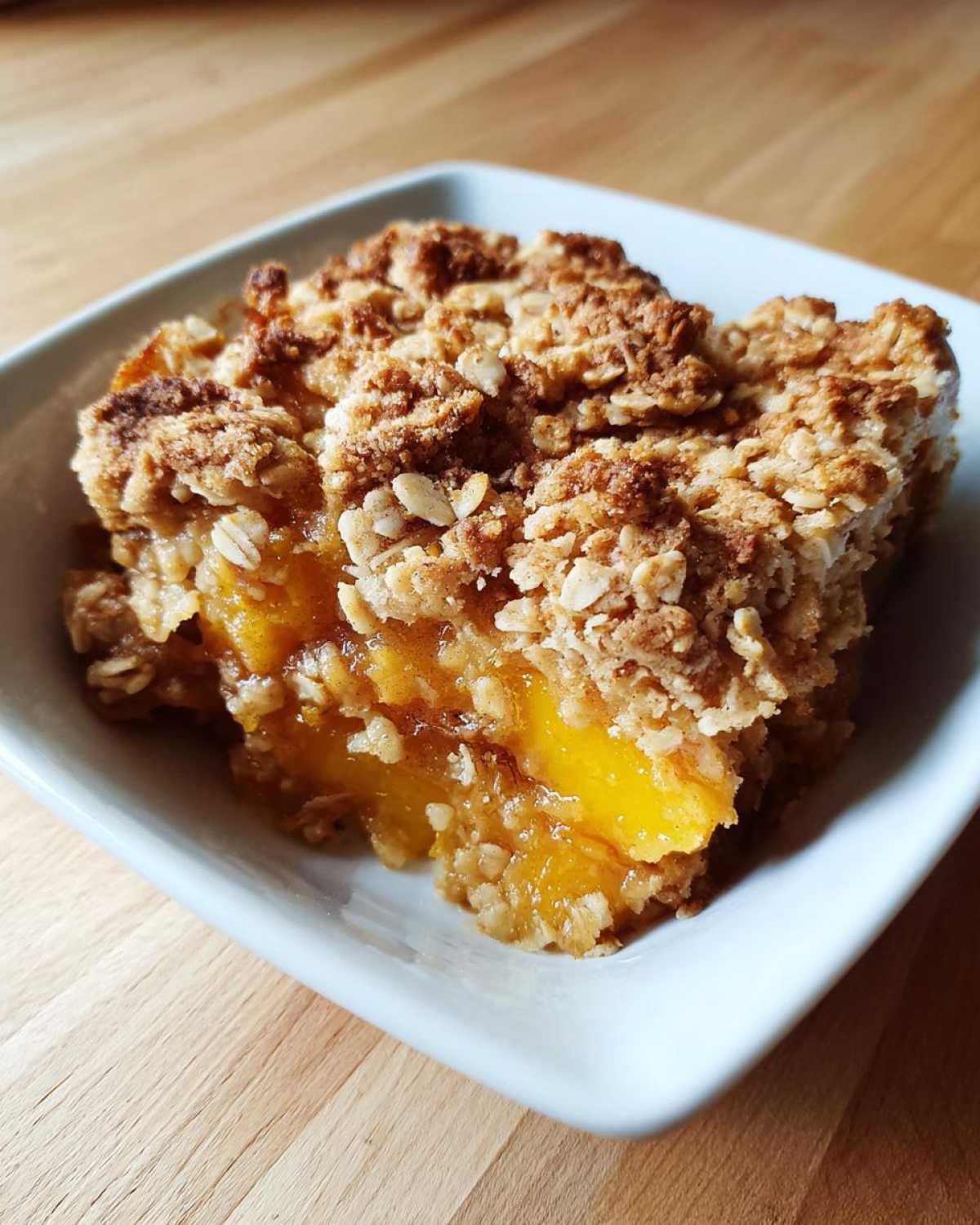A close-up of a square serving of warm Peach Crumble with a thick, golden oat topping in a white bowl.
