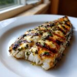 Close-up of a flaky, grilled tilapia fillet seasoned with spicy garlic herb marinade on a white plate.