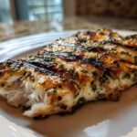 Close-up of a flaky, grilled tilapia fillet seasoned with herbs, showing distinct grill marks.