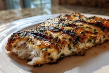 Close-up of a flaky, grilled tilapia fillet seasoned with herbs, showing distinct grill marks.