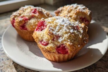Three freshly baked Strawberry Shortcake Muffins topped with streusel and powdered sugar, resting on a white plate.
