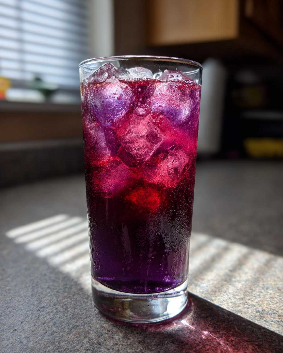 Tall glass filled with vibrant purple and pink Blueberry Lemonade over ice cubes, condensation visible.