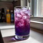 Close-up of a tall glass filled with colorful Blueberry Lemonade, ice cubes, and condensation.