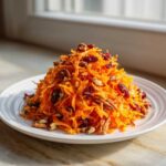 A mound of Zesty Apple Carrot Crunch Salad featuring shredded carrots, pecans, and dried cranberries on a white plate.